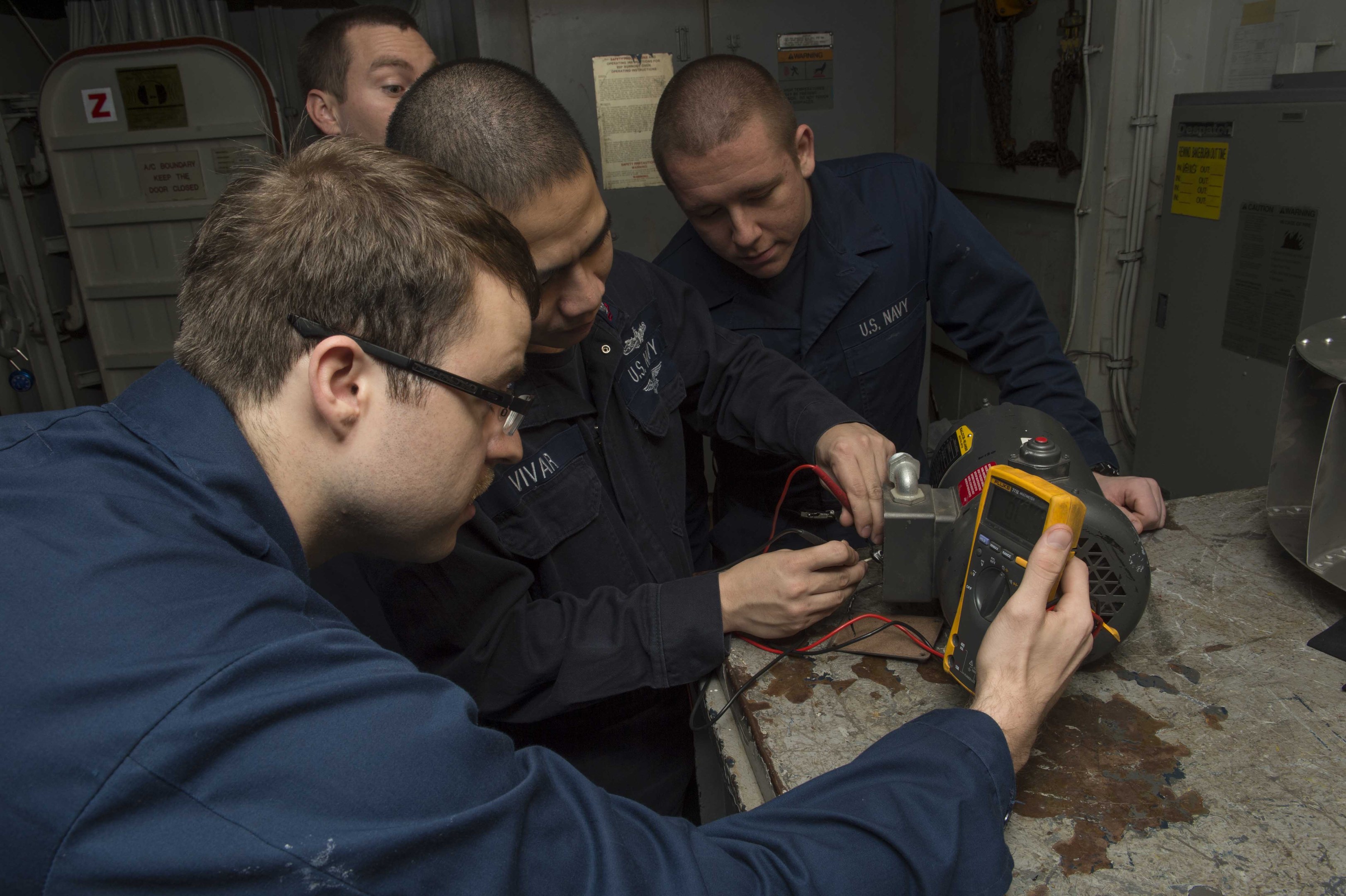 Vier Männer in einem Raum, die an einer Maschine arbeiten, mit einem Tisch voller Gegenstände auf der rechten Seite und Hintergrundmaschinen und Papieren an der Wand.