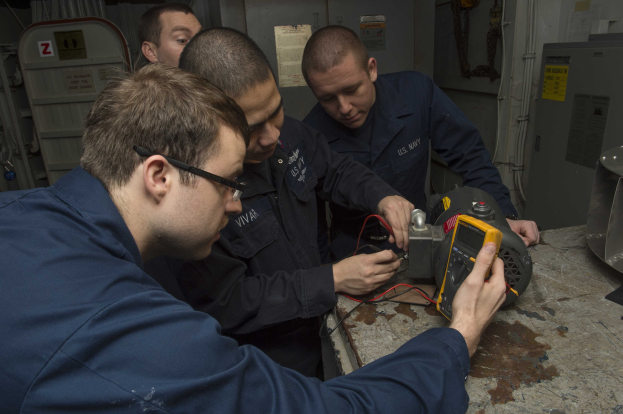 Vier Männer in einem Raum, die an einer Maschine arbeiten, mit einem Tisch voller Gegenstände auf der rechten Seite und Hintergrundmaschinen und Papieren an der Wand.