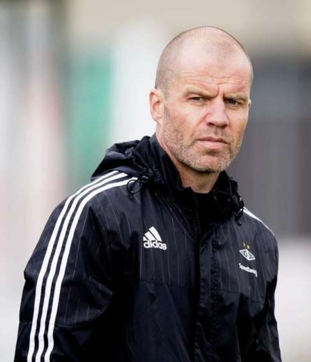 Baldiger Trainer in marineblauer Jacke mit weißen Streifen und weißem Kragen auf einem Fußballfeld mit gefalteten Händen und ernstem Gesichtsausdruck.