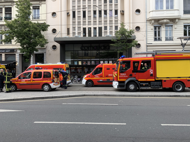 Eine Gruppe von Feuerwehrautos auf einer Straße in Paris geparkt, mit Menschen auf dem nahen Bürgersteig, Gebäuden, Bäumen und einem Fahrrad im Hintergrund.