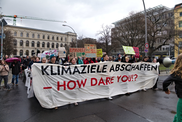 Eine Gruppe von Menschen marschiert mit einem Banner, auf dem "Klimaziele Abschaffen - Wie wagst du es?" steht, die Straße entlang, einige halten Schirme und einer verwendet ein Megafon, vor dem Hintergrund von Laternenmasten, Schildern, Bäumen, Gebäuden mit Fenstern, einem Kran und einem klaren blauen Himmel.