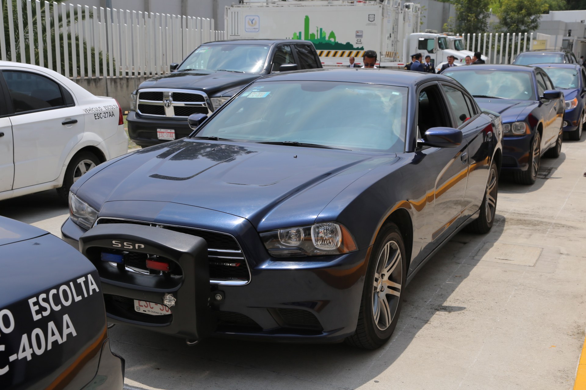 Polizeifahrzeuge auf einem Parkplatz mit Menschen auf der Straße, einem Zaun, Bäumen, Gebäuden und einem klaren Himmel im Hintergrund.