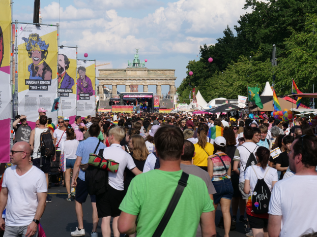 Eine große Menschenmenge marschiert vor dem Brandenburger Tor in Berlin, Deutschland, mit vielen, die Mützen und Taschen tragen, Schilder auf der linken Seite und Bäume, Zelte, Luftballons und ein Tor im Hintergrund unter einem bewölkten Himmel.