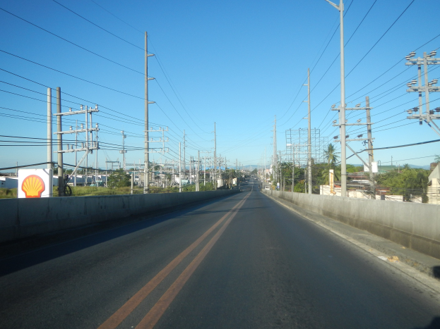 Autobahn mit Stromleitungen am Rand, flankiert von Mauern, Bäumen, Gebäuden und einem klaren blauen Himmel im Hintergrund.