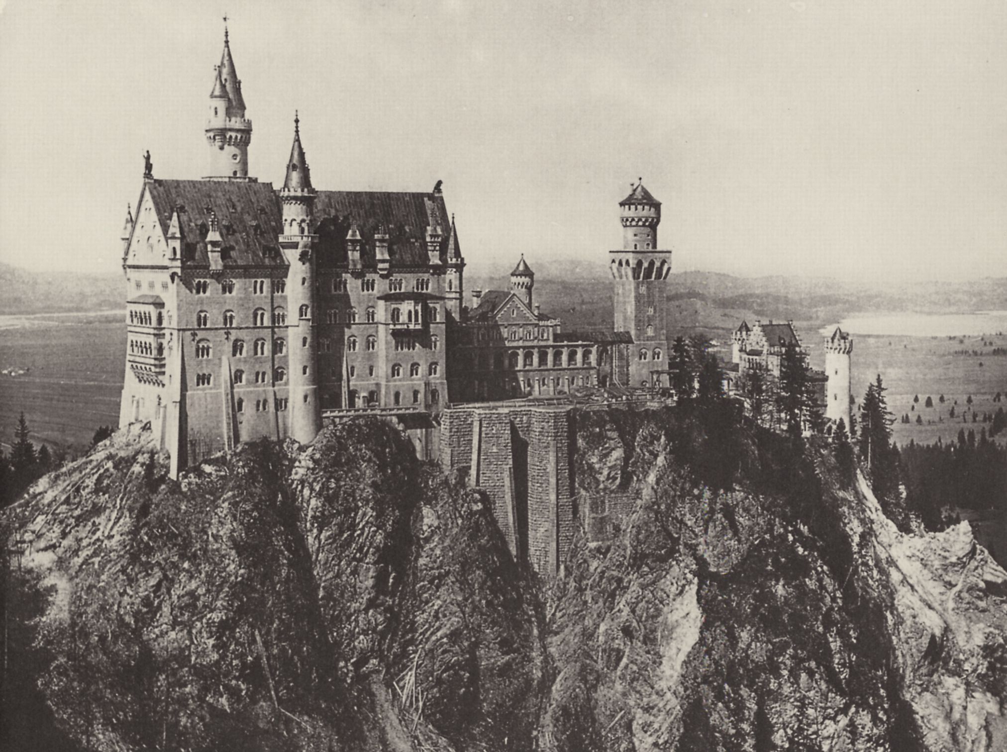 Schloss Neuschwanstein in Deutschland, thront auf einem Berg umgeben von Bäumen unter einem klaren Himmel, dargestellt in Schwarz-Weiß.