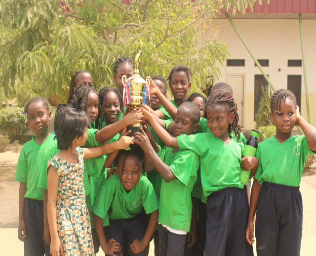 Eine Gruppe von Kindern hält einen Pokal in einem Schulhof mit Bäumen und einem Gebäude im Hintergrund.