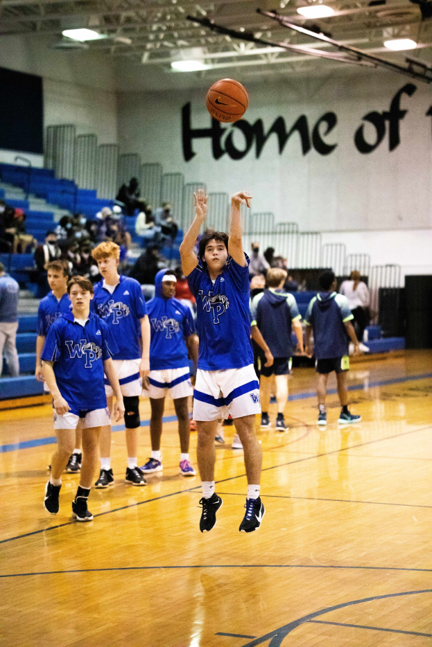 Eine Gruppe junger Männer, die Basketball auf einem Indoor-Court mit einer Wand spielen, die Text, Stufen, Geländer und Deckenleuchten sowie Personen mit Turnschuhen zeigt.