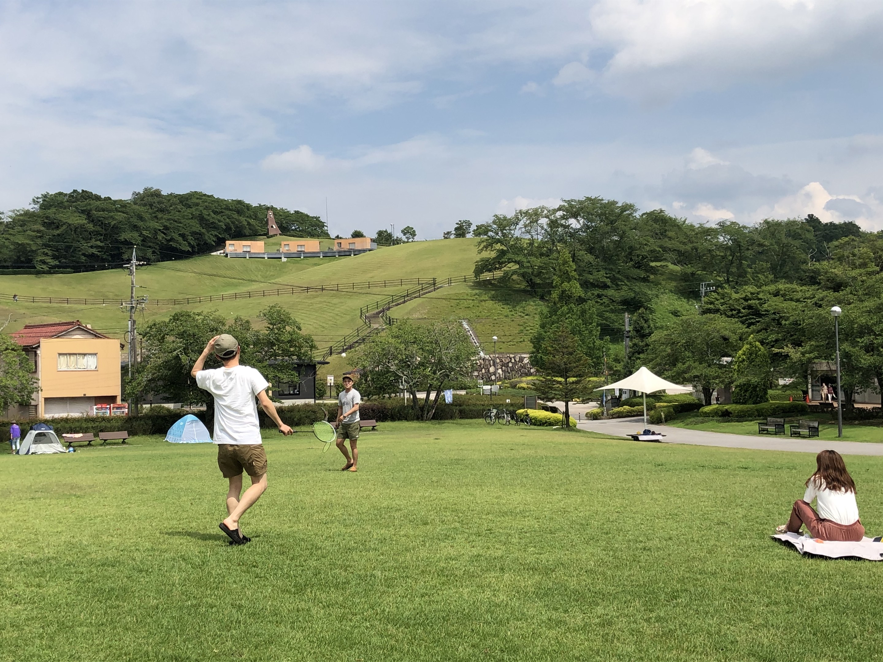 Gruppe von Menschen, die Badminton in einem Park spielen, mit einem Mann, der einen Schläger hält und auf einer Decke auf dem Gras sitzt, in der Nähe von Zelten und Gebäuden, mit Hügeln und bewölktem Himmel im Hintergrund.