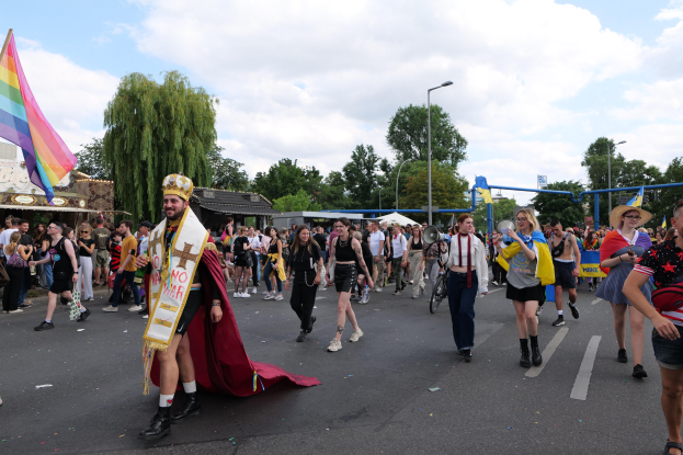 Eine Gruppe von Menschen marschiert bei der Pride Parade 2018, einige halten Musikinstrumente und tragen Mützen, während sie eine Regenbogenfahne eine Straße mit Laternen, Bäumen, Schuppen und einem bewölkten Himmel entlang tragen.