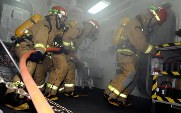 Eine Gruppe von Feuerwehrleuten in Schutzausrüstung, darunter Helme und Sauerstoffflaschen, arbeitet an einem Feuerwehrauto. Rechts ist ein Gestell mit verschiedenen Gegenständen zu sehen, und im Hintergrund gibt es Rohre, Lichter und andere Gegenstände.