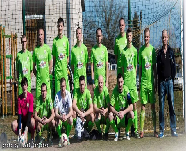 Gruppe von Männern in grünen T-Shirts auf einem Fußballfeld mit einem Ball vor ihnen und einem Torpfosten rechts daneben, Bäume und Gebäude im Hintergrund.