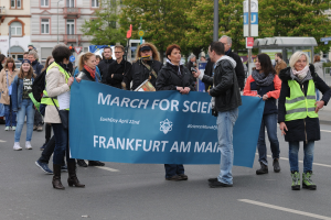 Gruppe von Menschen marschiert auf einer Straße mit einem "Marsch für die Wissenschaft Frankfurt am Main"-Schild mit Bäumen, Pfählen, Schildern, Gebäuden und einem klaren blauen Himmel im Hintergrund.