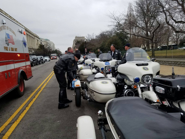 Eine Gruppe von Polizeibeamten steht neben Motorrädern am Straßenrand, mit einem Feuerwehrauto und anderen Fahrzeugen auf der Straße, Bäumen, Gebäuden und einem klaren blauen Himmel im Hintergrund.