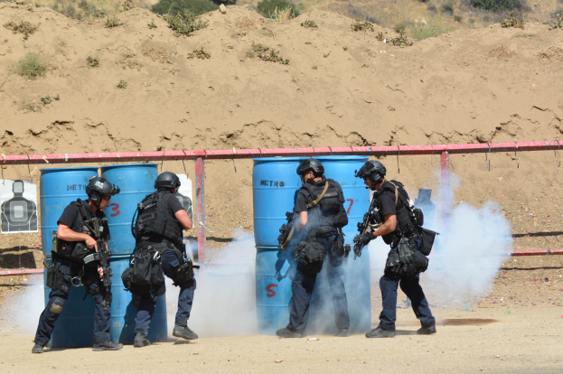 Eine Gruppe von Polizisten in schwarzer Einsatzkleidung, jeder mit einer Waffe in der Hand, steht auf einem Feld mit blauen Tonnen, einem roten Zaun, einem Plakat, Pflanzen und einem Berg im Hintergrund.