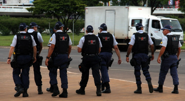 Eine Gruppe von Beamten in Uniformen, einschließlich Mötzen, Hemden, kugelsicheren Westen, Uhren, Hosen und Schuhen, die auf einer Straße mit einem geparkten Lkw rechts, Gebäuden, Bäumen, Pflanzen, Masten, Bannern und einer Brücke im Hintergrund gehen.