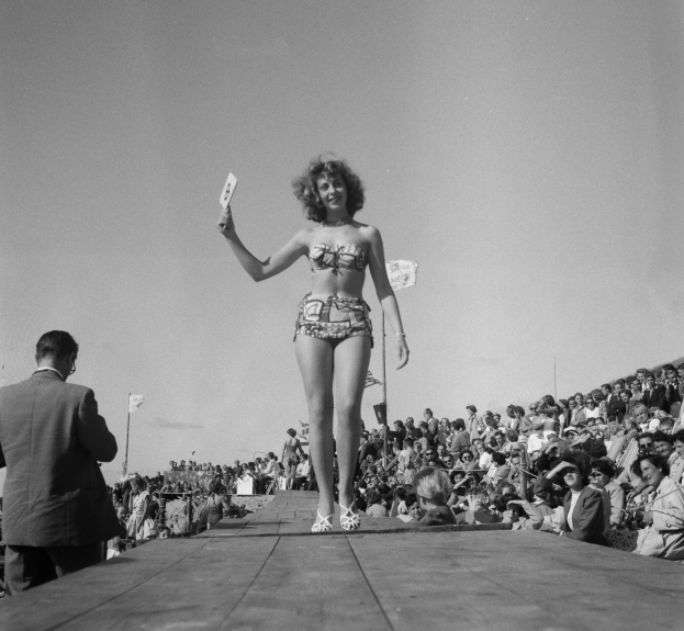Eine Frau in einem Bikini steht auf einer Bühne und hält eine Karte in der Hand, mit einem Mann zu ihrer Linken, vor einer Menge, unter einem sichtbaren Himmel, in einem Schwarz-Weiß-Bild.
