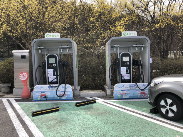 Ein Elektroauto auf der rechten Seite eines Parkplatzes neben einer Ladestation in der Mitte, mit Bäumen und einem klaren blauen Himmel im Hintergrund.
