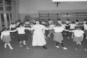 Schwarzes und weißes Foto von Kindern in Schuluniformen, die in einem Raum mit einem Kreuzsymbol an der Wand, Glastüren mit Vorhängen und Deckenleuchten tanzen.