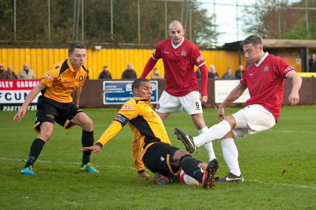 Spieler in blauen und roten Uniformen konkurrieren auf einem Rasenfeld mit einem Ball, während Zuschauer außerhalb des Spielfelds stehen und jubeln, mit Bäumen und Himmel im Hintergrund.