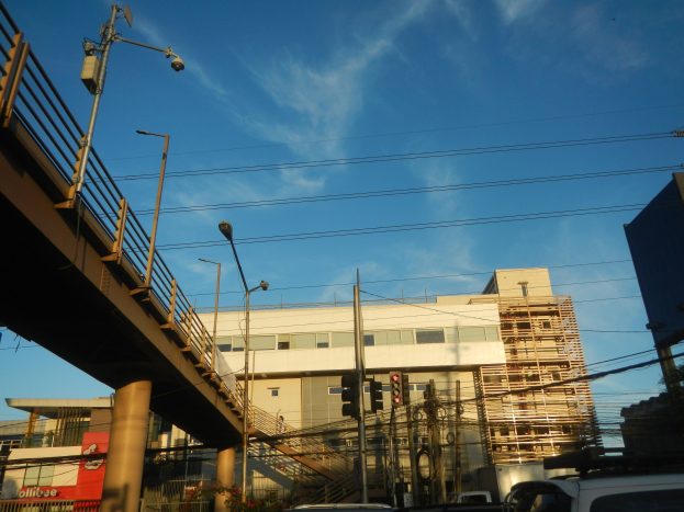 Stadtstraße mit parkenden Autos, Brücke, Laternen, Strommasten mit Kabeln, Ampeln, Gebäude mit Fenstern und bewölkter Himmel.