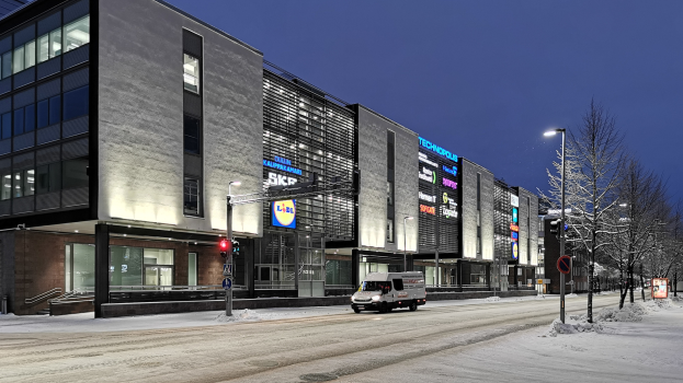 Ein Lieferwagen fährt nachts auf einer verschneiten Straße, umgeben von Gebäuden, Straßenlaternen, Verkehrspfosten, Verkehrszeichen, Schildern, Bäumen und einem sternenklaren Himmel.