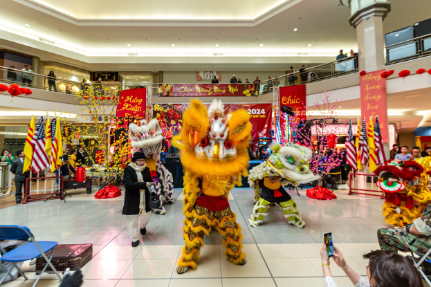 Ein lebendiges chinesisches Neujahrsfest in einem Einkaufszentrum mit Menschen in Kostümen, die tanzen, andere auf Stühlen sitzen und Handys halten, umgeben von Fahnen, Bannern, Dekorationen, Geländern, Säulen und Deckenleuchten.