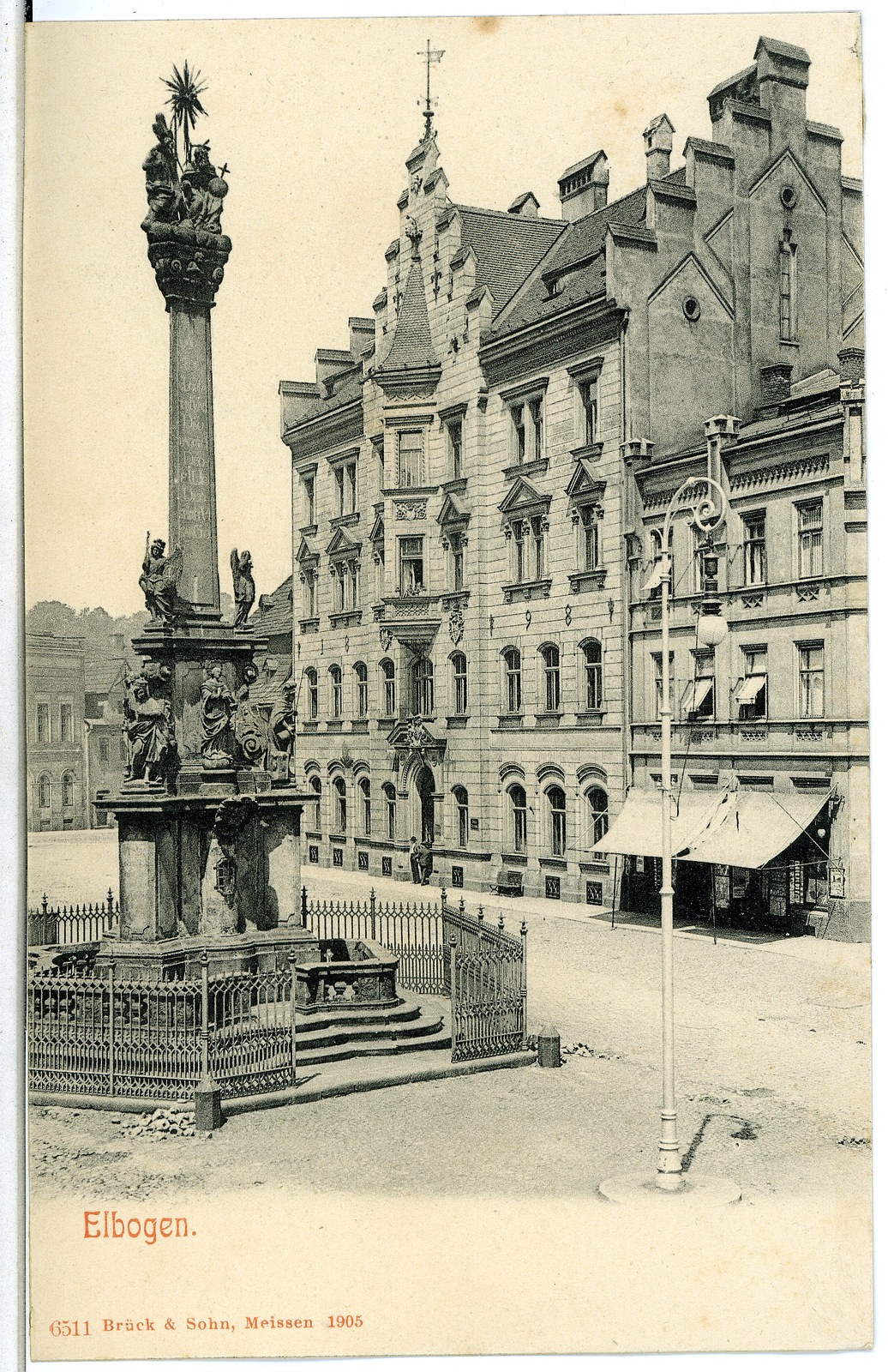 Ein altes Schwarz-Weiß-Foto eines Stadtplatzes mit einem Denkmal in der Mitte, umgeben von Gebäuden, Straßenlaternen, Gittern, Bäumen und einem klaren Himmel, mit Text am unteren Rand.