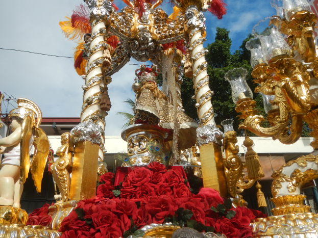 Ein Paradewagen mit roten Rosen und goldenen Verzierungen, darunter Statuen und Blumen, vor einem Gebäude, Bäumen und einem bewölkten Himmel.