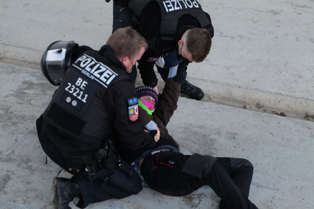 Ein Mann liegt am Boden mit zwei Polizeibeamten daneben, einer hält ein Kind; der Mann trägt eine Mütze und die Beamten haben Text auf ihren Jacken.