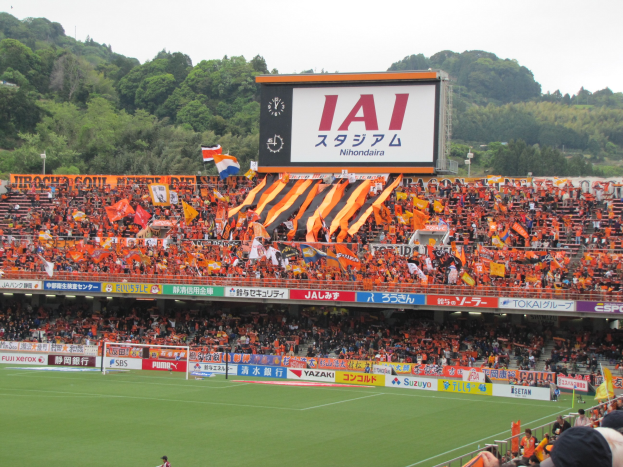 Ein Fußballspiel wird in einem Stadion mit einer großen Menschenmenge, grünem Rasen, einem Tor, Bannern, Fahnen, einem großen Bildschirm, Bäumen und einem klaren blauen Himmel gespielt.