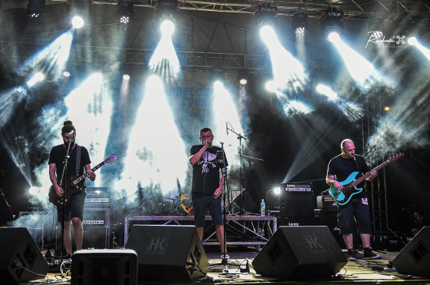 Drei M├Ąnner auf der B├╝hne, die Gitarren spielen und in Mikrofone singen, umgeben von Musikausr├╝stung und einem Banner mit Text und Lichtern, auf dem Belgrade Music Festival in Serbien im Jahr 2016.