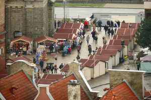 Eine belebte Stadtstraße mit Fußgängern, Gebäuden, Ständen, einem Baum und einer Bahnstrecke unter einem bewölkten Himmel.