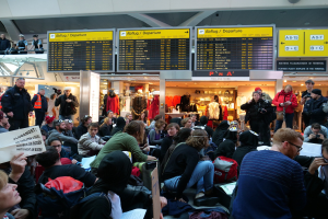 Eine große Gruppe von Menschen in einem Flughafen, einige sitzen mit Taschen und Papieren, andere stehen, mit Texttafeln, Schaufensterpuppen in Kleidern und Deckenbeleuchtung im Hintergrund, was auf eine Protestveranstaltung hindeutet.