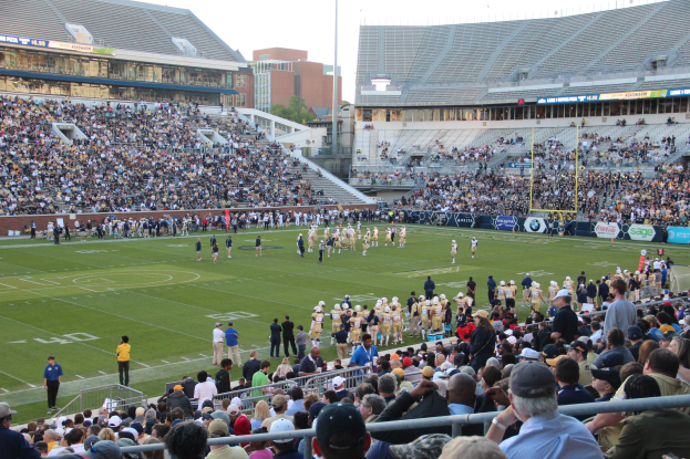 Ein Fußballspiel im Stadion mit Spielern auf dem Feld und Zuschauern in den Rängen unter einem klaren blauen Himmel.
