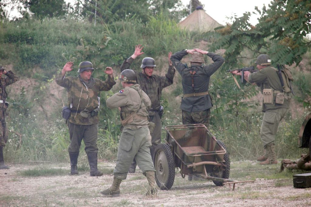 Mehrere Personen in militärischer Uniform und Helmen stehen neben einem Wagen, umgeben von Pflanzen, Bäumen und Gras unter einem klaren Himmel.