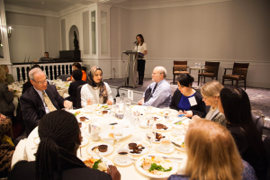 Eine Gruppe von Menschen sitzt um einen Esstisch mit Tellern, Besteck und Trinkgefäßen, während eine Frau in der Nähe eines Podiums mit einem Mikrofon im Hintergrund steht.