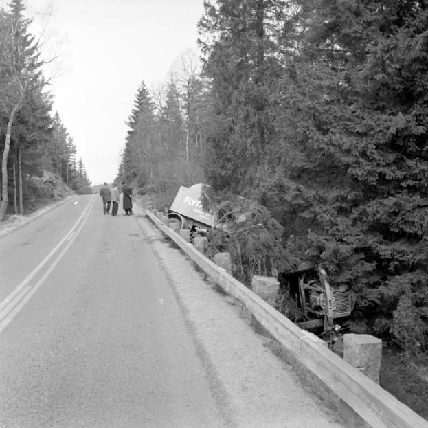 Ein Schwarz-Weiß-Foto eines Lastwagens, der auf der Seite der Straße gegen einen Baum geprallt ist, mit ein paar Menschen auf der linken Seite und vielen Bäumen im Hintergrund.