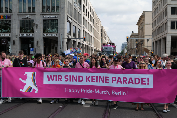 Eine Gruppe von Menschen marschiert auf einer Straße in Berlin, Deutschland, mit einer pinken Fahne, auf der "Happy Pride March" steht, vorbei an Gebäuden, Laternenpfählen und Verkehrsampeln unter einem bewölkten Himmel.