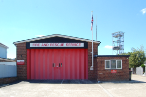 Feuerwehr- und Rettungsdienstgebäude mit roter Tür, Fenstern, einem Namensschild, einem Fahnenmast mit Flagge, einem Metallturm, einem Zaun, einer Baumgruppe und einem bewölkten Himmel.