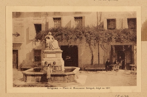 Ein altes Foto eines Brunnens mit einer Statue auf einem Sockel in der Mitte, umgeben von Menschen, die auf Bänken sitzen, mit einem Baum im Hintergrund und Text unten.