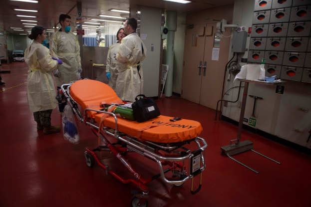 Eine Gruppe von Menschen in Schutzausrüstung steht um eine Trage in einem Krankenhauszimmer, das mit medizinischer Ausrüstung gefüllt ist, darunter ein Stretcher mit einer Tasche und anderen Gegenständen, ein Ständer mit verschiedenen Gegenständen, eine Tür, eine Säule, ein Schild und eine Decke mit Lampen.