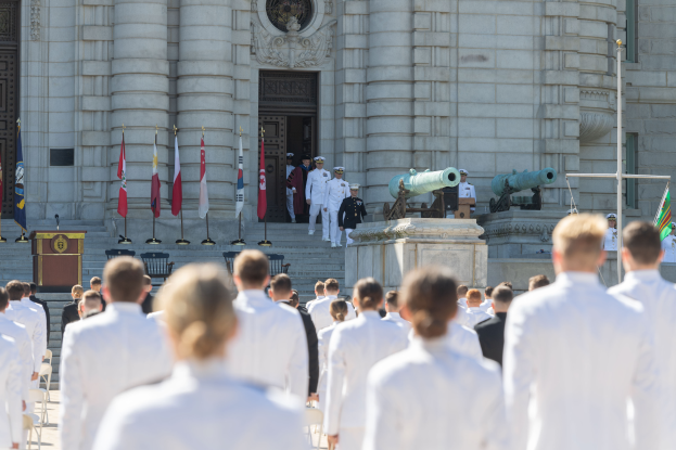 Eine Gruppe von Menschen in weißen Uniformen und Mützen steht vor einem Gebäude mit Säulen und einer Tür, mit Fahnen, einem Podium mit Mikrofon, Stufen und Kanonen im Hintergrund, was auf eine Abschlusszeremonie an einer Marineakademie hindeutet.