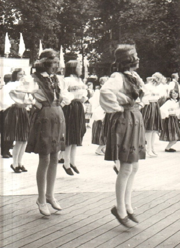 Eine Gruppe von Frauen in traditioneller bayrischer Tracht tanzt auf einem Holzboden umgeben von Bäumen und Fahnen, dargestellt in Schwarz-Weiß.