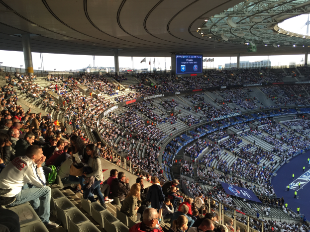 Eine große Menschenmenge sitzt im Allianz Stadion in München, Deutschland, und schaut ein Fußballspiel, mit einer Bühne rechts, Fahnen, Stangen und einem Bildschirm im Hintergrund und dem Himmel oben sichtbar.