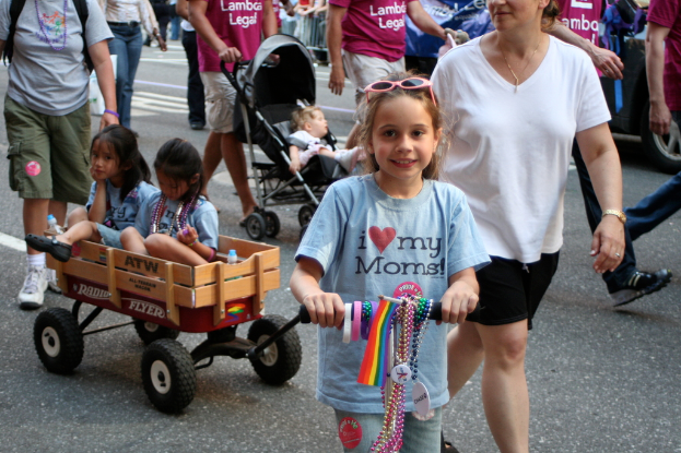 Eine Gruppe von Menschen, darunter Mädchen und ein Kinderwagen mit einem Kind, geht auf einer Straße mit einer Mauer und einem Fahrzeug im Hintergrund.