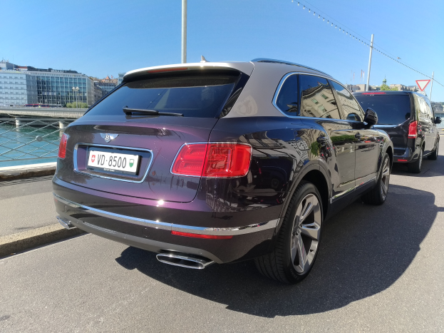 Ein Bentley Bentayga V8 S, der auf einer Straße mit einer Brücke, Gebäuden, Bäumen und einem klaren blauen Himmel im Hintergrund fährt.