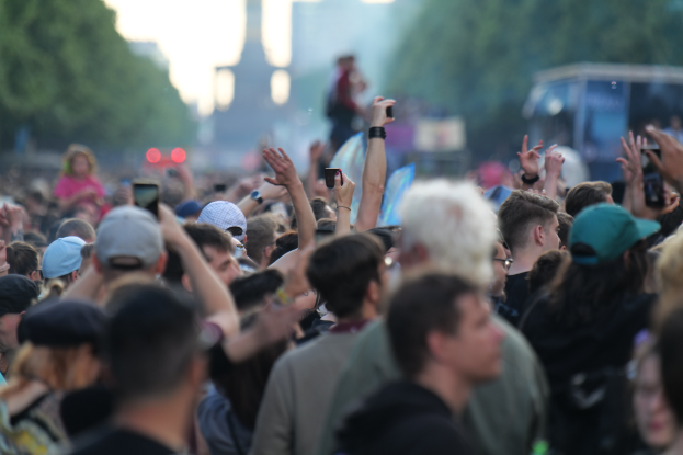 Menschenmenge auf einem Musikfestival mit erhobenen Händen, einige tragen Mützen und halten Handys, im Hintergrund leicht unscharfe Bäume, Gebäude, Lichter und ein Fahrzeug.