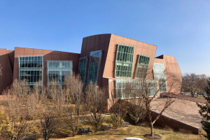 Großes mehrstöckiges Gebäude mit zahlreichen Fenstern, umgeben von Bäumen, Bänken, Gras und einem Laternenmast, mit anderen Gebäuden und einem klaren blauen Himmel im Hintergrund, identifiziert als der neue Campus der University of Illinois at Chicago.