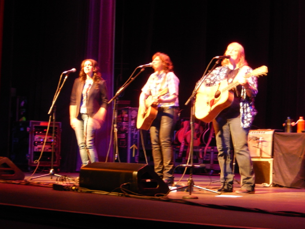Drei Frauen performen auf der Bühne eines Musikkonzerts, singen in ein Mikrofon und spielen eine Gitarre, mit einem Tisch voller Wasserflaschen und Gitarren dahinter.