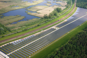 Eine Luftaufnahme einer Solaranlage mit Panelen in einem Feld, umgeben von Bäumen, Gras, Pflanzen und Wasser, mit einem Zug auf einem Bahngeleis in der Nähe.
